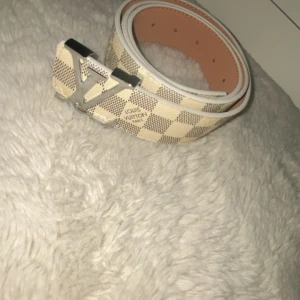 Louis Vuitton rutigt bälte med LV-spänne - Snyggt bälte från Louis Vuitton med klassiskt rutigt mönster i beige och brunt. Bältet har en stor, silverfärgad LV-logga som spänne och vita kanter. Perfekt accessoar för att lyfta din outfit med en lyxig touch.