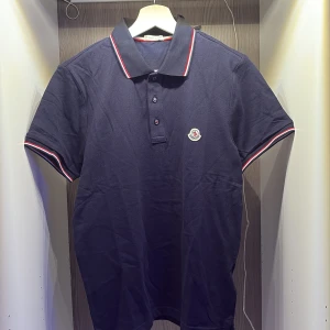 Mörkblå Moncler pikétröja - Snygg mörkblå pikétröja från Moncler med klassisk krage och rödvit rand längs krage och ärmslut. Broderad Moncler-logga på bröstet och tre knappar framtill. Tillverkad i bomull för en skön och stilren känsla. Pris går alltid att diskutera!!