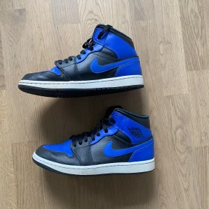 Nike Air Jordan 1 svart/blå sneakers - Snygga Nike Air Jordan 1 sneakers i svart och blått skinn med klassisk vit sula och svarta skosnören. Ikonisk design med högt skaft och tydlig Nike swoosh på sidan. Perfekta för dig som vill sticka ut med en streetstyle-look.