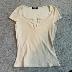 Topp med v-ringning  - Snygg beige topp från Fashionkilla med korta ärmar och unik v-ringning framtill. Modellen är figurnära och har en enkel, stilren design som passar perfekt till jeans eller kjol. Mjuk och stretchig kvalité för en bekväm känsla.