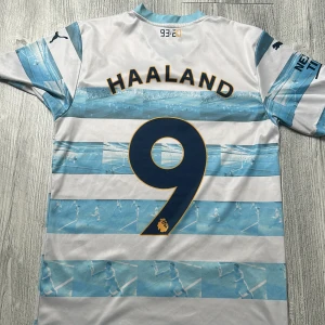 Manchester City fotbollströja Puma 176 - Agueros last minute goal. Manchester City matchtröja från Puma i vitt och ljusblått med randigt mönster och klubbmärke på bröstet. Tröjan har korta ärmar, V-ringning med ljusblå kant och Etihad Airways-tryck. Materialet är lätt och andas, perfekt för fotbollsträning. Agueros last minute goal