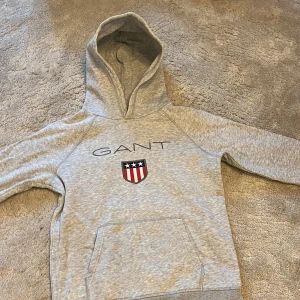 Grå hoodie från GANT med logga - Snygg grå hoodie från GANT med klassisk logga och sköld på bröstet. Tröjan har huva, magficka och långa ärmar. Perfekt för en chill och sportig look. Mjuk och skön i bomullsmix.