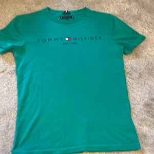 Grön t-shirt från Tommy Hilfiger - Snygg grön t-shirt från Tommy Hilfiger med tryckt logga och text på bröstet. Tillverkad i mjuk ekologisk bomull, klassisk passform och rund halsringning. Perfekt för dig som gillar stilrena och bekväma plagg med en touch av premiumkänsla.