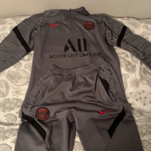 Grå PSG träningsset från Nike - Snyggt grått träningsset från PSG och Nike med svarta detaljer och röda loggor. Tröjan har halv dragkedja och byxorna har resår i midjan. Perfekt för dig som älskar fotboll och vill ha en sportig look.