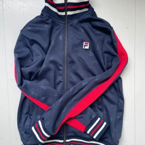 FILA tracktop jacka nyskick - Snygg blå track jacket från FILA med röda ränder längs ärmarna och vita/röda detaljer på muddar och krage. Nyskick, bara provad hemma. Jag är 178 och den sitter bra och relaxed på mig. Den är en Small men passar absolut Medium också. Jackan har dragkedja framtill, diskret mönster i tyget och klassisk FILA-logga på bröstet. Perfekt för en sportig och casual vibe i sommar.  