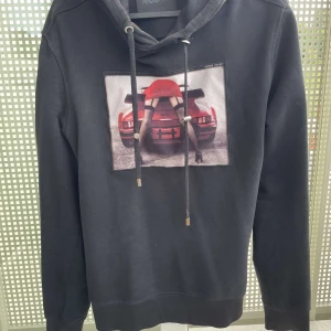Svart hoodie Limitato x Riocam med print - Svart hoodie från Limitato x Riocam med stor grafisk print framtill som visar en röd sportbil och en person i röd kjol. Tröjan har huva med snörning och långa ärmar. Perfekt för dig som gillar streetwear med attityd.