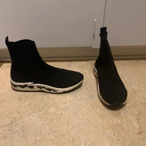 Svarta sock-sneakers Sandro Paris  - Svarta sock-sneakers med högt skaft och elastisk ovandel i stickad syntet. Sulan är vit med svarta detaljer i vågigt mönster, vilket ger en cool och modern look. Perfekta för dig som gillar streetstyle och vill ha något bekvämt och trendigt.