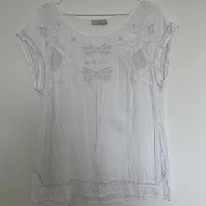Vit topp med broderade detaljer från Cream - Säljer en  vit topp från Cream med broderade detaljer och utskurna mönster framtill. Blusen har korta ärmar och rund halsringning. Perfekt för varma dagar och ger en bohemisk känsla.