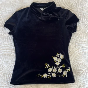 Svart sammetsblus med broderade blommor från Einde - Säljer en svart sammetsblus från Einde med korta ärmar och unik krage. Nedtill finns fina broderade vita och gula blommor som ger toppen en söt touch. Perfekt för dig som gillar detaljer och mjuka material.