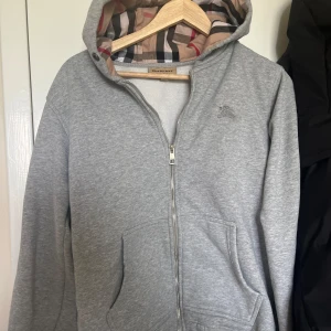 Grå hoodie med dragkedja från Burberry - LÄGG BUD!!! Säljer en grå hoodie från Burberry med dragkedja framtill och klassiskt rutigt foder i huvan. Tröjan har två fickor och en diskret Burberry-logga på bröstet. Perfekt för en stilren och bekväm look. 