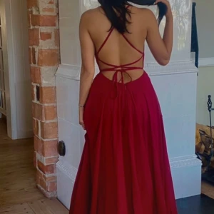 Vinröd långklänning med öppen rygg och slits - Säljer en vinröd långklänning med smala axelband som korsas i ryggen och knyts baktill. Klänningen har en elegant slits framtill och en öppen rygg, vilket ger en snygg och modern look. Perfekt för fest eller bal!