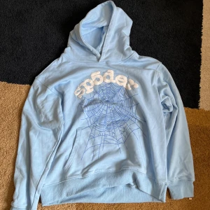 AU! Ljusblå hoodie från spyder -  Hör av dig innan du köper! Säljer en ljusblå hoodie från Spyder med stort vitt tryck och spindelnät på bröstet. Texten är dekorerad med små glittriga stenar runtom. Mjuk bomullskvalitet och klassisk huva. Perfekt för dig som gillar streetwear och unika detaljer.