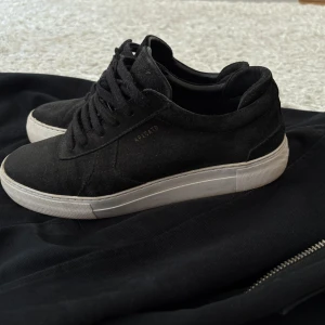 Svarta sneakers från Axel Arigato - Säljer ett par svarta sneakers från Axel Arigato med vit platt sula och snörning. Skorna har en stilren design i mocka och passar perfekt till en avslappnad look. Märket syns diskret på sidan och sulan.