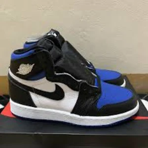 Nike Air Jordan 1 Retro High OG 'Game Royal' - Nike Air Jordan 1 Retro High OG i färgerna svart, vit och blå. Klassisk high-top sneaker med svart swoosh, vit bas och blå detaljer på tå och häl. Skon har snörning och rund tå, perfekt för dig som gillar streetwear och sneakers. Jag köpte dom från USA så det blev extra dyrt!!