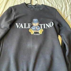 Svart sweatshirt från Valentino Paris - Svart sweatshirt från Valentino Paris med tryck framtill av en björn i blå kläder och stor vit logga. Rund halsringning och långa ärmar. Perfekt för en stilren och trendig look.