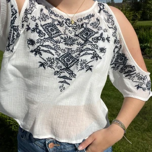 Broderad vit blus  - Snygg vit blus från H&M Coachella med mörkblått broderat mönster. Blusen har öppna axlar och knytband i nacken med tofsdetaljer. 