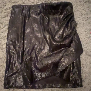 Svart paljettkjol från Pimkie - Säljer en svart glittrig paljettkjol från Pimkie. Kjolen är kort och har en rak passform med snygg omlott-detalj framtill. Perfekt för dig som vill sticka ut med lite extra glitter.