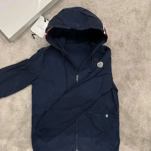 Blå vindjacka från Moncler (Med kvitto) - Säljer en mörkblå vindjacka från Moncler med huva och dragkedja. Jackan har Moncler-logga på ärmen och snygga rödvita detaljer på huvan. Perfekt för blåsiga dagar och enkel att matcha med det mesta.  NFC funkar, tags och sådant medföljer. Den är i nytt skick, använd ett fåtal gånger. Säljs då den inte kommer till användning 