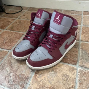 Nike Air Jordan 1 Mid cherrywood red - Säljer ett par Nike Air Jordan 1 Mid i färgerna cherrywood red och grå. Skorna har klassisk hög siluett, vit yttersula och bordeauxfärgade detaljer med snörning. Perfekta för dig som gillar streetwear och sneakers med ikonisk design.