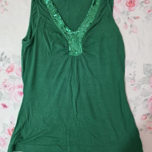 Grön ärmlös topp med paljettdetalj - Snygg grön ärmlös topp med djup v-ringning dekorerad med gröna paljetter. Perfekt för att piffa upp din outfit med lite extra glans. Mjuk och skön att bära.