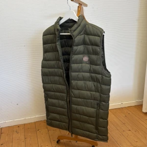 Olivgrön dunväst från Kronstadt - Snygg olivgrön dunväst från Kronstadt med quiltad design och dragkedja framtill. Västen har en hög krage och två fickor med dragkedja. Perfekt för lager-på-lager och enkel att matcha med olika outfits.