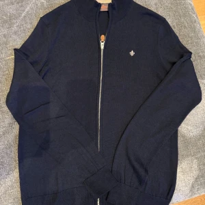 Mörkblå ziptröja från Morris - Säljer en mörkblå ziptröja från Morris i 100% merinoull. Tröjan har en liten broderad logga på bröstet, hög krage och dragkedja framtill. Perfekt för lager-på-lager och stilren look.