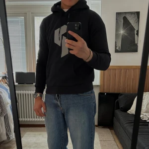 Svart hoodie från Peak Performance - Säljer en svart hoodie från Peak Performance med stor grå logga på bröstet. Tröjan har huva med dragsko och en stor magficka. Perfekt för en avslappnad stil och enkel att matcha med jeans eller joggers.