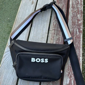 Svart magväska från BOSS - Säljer en svart magväska från BOSS med vit logga framtill. Väskan har ett stort huvudfack med dragkedja och ett mindre ytterfack och ett till fack på insidan. Justerbart band i svart, vitt och beige. Perfekt för att bära det viktigaste med stil.