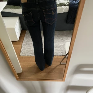 Mörkblå jeans med färgglada sömmar - Säljer ett par mörkblå jeans med raka ben och snygga kontrastsömmar i rosa. Byxorna har låg midja och bakfickor med lock och knappdetalj. Perfekta för dig som gillar klassiska jeans med en twist.