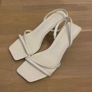 Vita sandaler med klack och fyrkantig tå - Säljer ett par stilrena vita klackar med smala remmar och fyrkantig tå. Skorna har öppen häl och en elegant klack som ger en snygg siluett. Perfekta för dig som gillar minimalistisk design och vill ha något fräscht till sommaren.