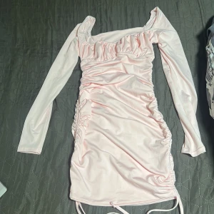 Ljusrosa långärmad omlottklänning - Säljer en söt ljusrosa kortklänning med långa ärmar och rynkade detaljer längs sidorna. Klänningen har omlottkänsla och en lätt glansig finish som ger en elegant look. Perfekt för dig som gillar feminina och söta klänningar till sommaren.