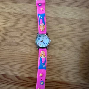 Rosa barnklocka med sjöjungfrumotiv - Säljer en färgglad klocka med rosa armband och motiv av sjöjungfru och stjärnor. Urtavlan har tydliga siffror i olika färger och silverfärgat hölje. Perfekt accessoar för den som gillar lekfull stil.