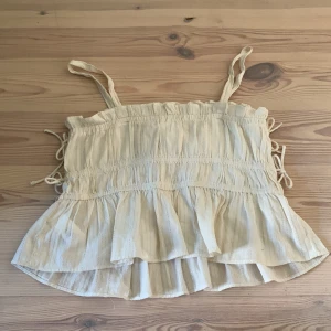 Beige volangtopp med smala axelband från Zara - Supersöt beige topp från Zara med smala axelband och volang nertill. Toppen har rynkade detaljer och knyt i sidorna. Perfekt för varma dagar och ger en luftig känsla.