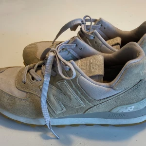 Beige sneakers från New Balance - Snygga beige sneakers från New Balance med klassisk design. Skorna har ovandel i mocka och textil, snörning och vit sula med gummidetaljer. Perfekta för en avslappnad och sportig stil.
