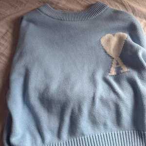 Ljusblå stickad tröja från Ami Paris - Säljer en ljusblå stickad tröja från Ami Paris med deras ikoniska hjärta och A-logga i vitt på bröstet. Tröjan har rund halsringning och ribbade muddar. Perfekt för dig som gillar stilrena och trendiga plagg.