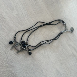 Svart halsband med hjärta och vingar - Unikt halsband med svarta band, dekorerat med svarta pärlor, silverfärgade detaljer och ett hängsmycke i form av ett hjärta med vingar. Justerbar längd med kedja och lås. Perfekt för att ge din outfit en cool touch.