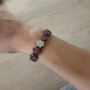 Lila armband med blommor - Säljer ett snyggt armband  med lila blommor i glittrande stenar och en silverfärgad blomformad detalj med logga. Perfekt accessoar för att lyfta din outfit med lite extra färg och bling.