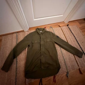 Olivgrön overshirt - Säljer en olivgrön overshirt från med två bröstfickor och patchdetaljer på ärmen. Jackan har klassisk krage och stängs med dragkedja och knappar. Perfekt att bära som lager över t-shirt eller skjorta. Priset är inte hugget i sten 👌
