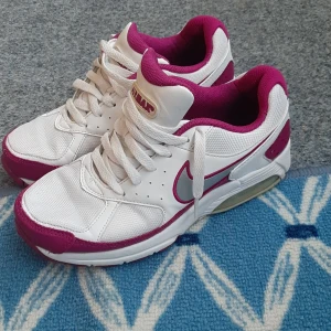Nike Air Max sneakers i vitt och rosa - Säljer ett par Nike Air Max sneakers i vitt med rosa detaljer och logga. Skorna har en klassisk sportig design med snörning och synlig Air Max-dämpning i sulan. Perfekta för dig som gillar färg och vill ha bekväma sneakers till vardags.