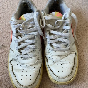 Vita Nike sneakers med rosa swoosh - Nike sneakers i vitt med en tydlig rosa swoosh på sidan. Skorna har vit snörning, perforerade detaljer på tån och svart sula. Låg modell med klassisk design och retrovibbar. Passar dig som gillar sportig och stilren look.