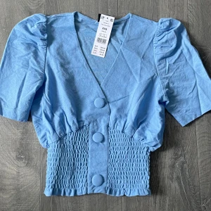 Ljusblå croppad blus med puffärm - Säljer en ljusblå croppad blus från Gina Tricot med v-ringning, puffärmar och smockad midja. Blusen har stora dekorativa knappar framtill och korta ärmar. Perfekt för dig som vill ha en trendig och söt topp! Prislappen är kvar, då den aldrig kommit till användning!