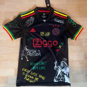 Ajax x Bob Marley svart fotbollströja - Säljer en svart Ajax fotbollströja från Adidas med Bob Marley-tema. Tröjan har färgglada ränder i rött, gult och grönt på axlarna, tryck med Ziggo-logga, Ajax-märke och flera Bob Marley-citat och illustrationer. Perfekt för dig som gillar både fotboll och reggae. ✅Mer tröjor hittas i profilen✅