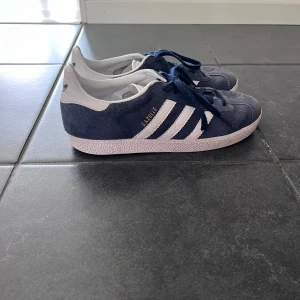 Adidas Gazelle i blå mocka - Säljer ett par klassiska Adidas Gazelle sneakers i blå mocka med vita detaljer och snörning. Skorna har de ikoniska tre ränderna på sidan och Adidas-logga på baksidan. Använda lite grann men fortfarande bra skick