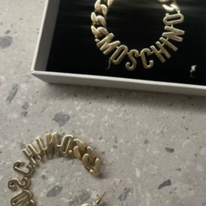 Guldiga örhängen från H&M x Moschino - Säljer ett par stora, runda örhängen i guldmetall från H&M x Moschino. Örhängena har en chunky kedjedesign och bokstäver som bildar 'MOSCHINO'. Perfekta för att sticka ut och ge en cool statement-look.