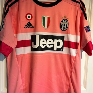 Juventus Pogba 10 - Juventus bortatröja i rosa med svarta och röda detaljer, Adidas-logga och Jeep sponsortryck. Pogba 10 på ryggen. Utrustad med Champions League och UEFA Respect patchar på ärmarna. Rund hals och korta ärmar.