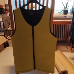 Gul väst med svarta detaljer - Säljer en gul sportväst med svarta kanter och dragkedja framtill. Västen är ärmlös och har en enkel, sportig design. Materialet är neopren och polyester vilket gör den lätt och smidig att bära. Storlek large 45*61 cm