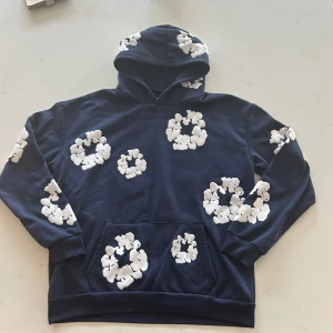 Mörkblå hoodie med vita blommor från Denim Tears - Säljer en mörkblå hoodie från Denim Tears med vita blommor tryckta över hela tröjan, inklusive huvan och fickan. Hoodien har en klassisk känguruficka och en skön passform. Perfekt för dig som vill sticka ut med en unik streetwear-stil. Pris kan diskuteras och skickar oftast inom 24 timmar