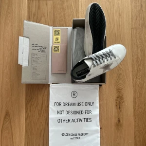  Golden Goose  - Golden Goose sneakers i vitt skinn med grå snörning och en grå stjärna på sidan. Skorna har svart hälparti och klassisk platt sula. Levereras i originalkartong med dustbag.