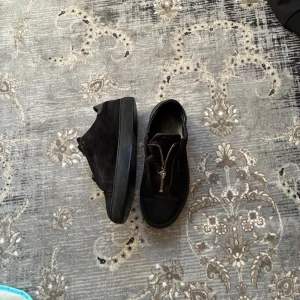 Axel arigato - Svarta sneakers Axel arigato i mocka med dragkedja framtill istället för klassisk snörning. Skorna har rund tå och platt sula, vilket ger en stilren och modern look. Perfekta för dig som vill ha något enkelt men ändå snyggt.