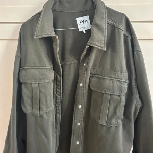 Olivgrön overshirt/jeansjacka från ZARA - Säljer en olivgrön overshirt/jeansjacka från ZARA med klassisk krage och stora bröstfickor med lock. Jackan har långa ärmar med manschett och knappar framtill. Perfekt att styla lager på lager för en cool look.